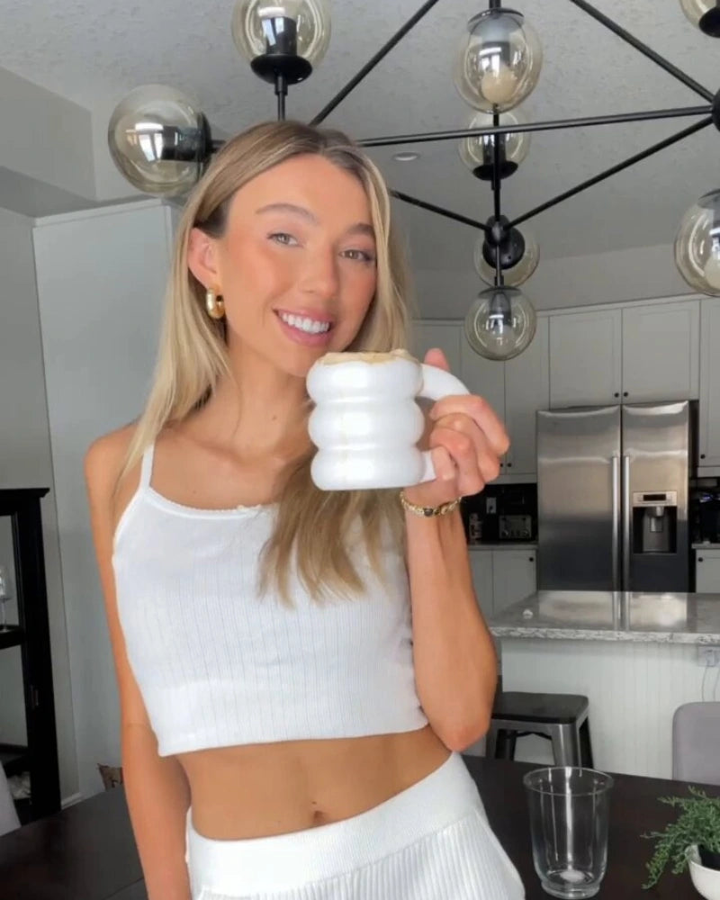 Woman smiling and holding a white mug, standing in a modern kitchen with a black chandelier and silver appliances. She is wearing a white tank top and pants.